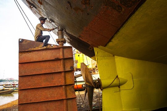 Dhaka, Bangladesh - 20 April 2026: View of a worker painting a ship's hull, contrasting vibrant yellow against dark, weathered rust and industrial machinery.
