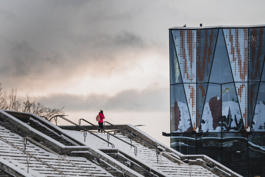View of a solitary figure in vibrant pink ascends snow-dusted steps towards a striking modern glass building under a moody sky, Tallinn, Estonia.