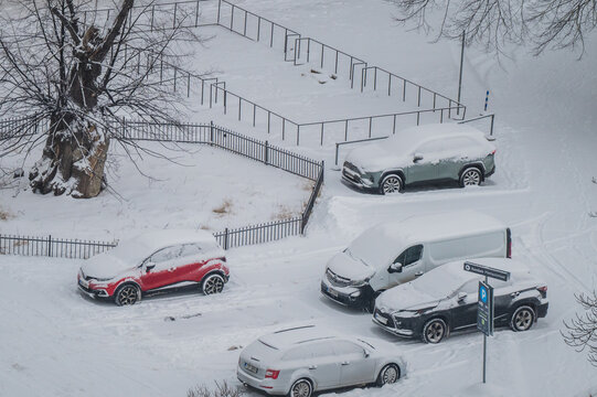 Tallinn, Estonia - 03 January 2025: View of snow blanketing a quiet urban scene, where vibrant cars peek through thick white layers beside skeletal trees and dark fences.