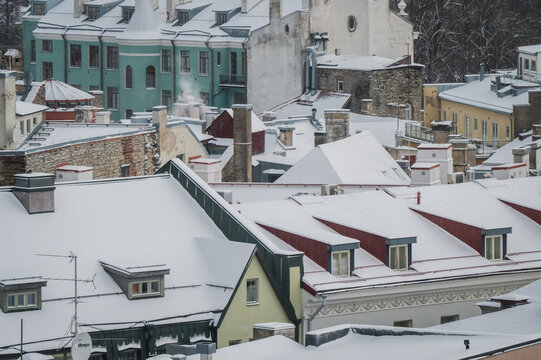 View of a snow-dusted cityscape with rooftops adorned in white blankets contrasting against the muted pastel buildings, smoke rising from a chimney, Tallinn, Estonia.