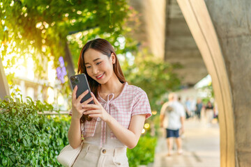 Fototapeta premium Young Asian woman reading a smartphone screen under an elevated path beside a hedge