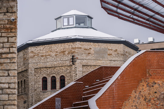 View of a historic stone tower with a snow-dusted roof stands beside a brick staircase under a gray sky, architecture blending old and new, Tallinn, Estonia.