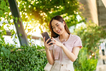 Fototapeta premium Young Asian woman smiling at a smartphone beside a hedge beneath a concrete overpass