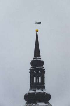 View of a dark spire reaching into the overcast winter sky, topped with a golden ball and weather vane dusted with snow, Tallinn, Estonia.