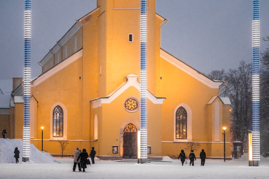 View of a brightly illuminated yellow church contrasting against the snowy ground, framed by modern light installations, creating a blend of history and innovation, Tallinn, Estonia.