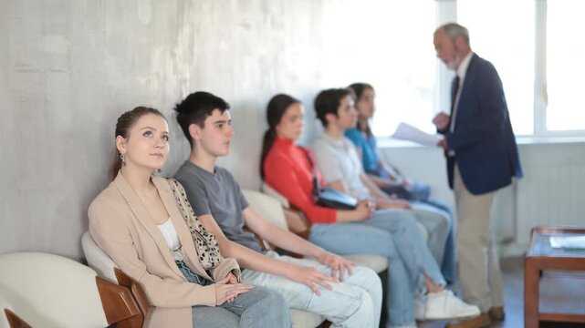 Relaxed young visitors sitting in a queue while senior manager speaking to one in background