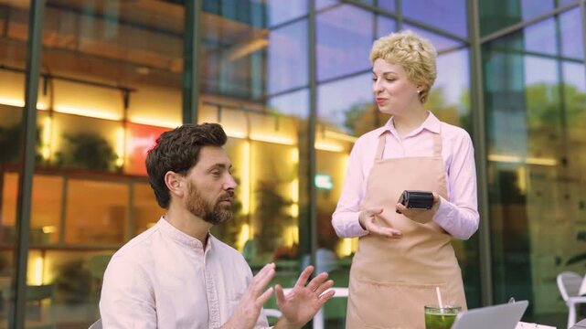 A man uses his smartphone to make a contactless payment to a waitress holding a card reader outside a modern restaurant