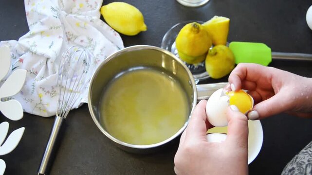 Close-up of hands cracking an egg into a bowl with visible yolk and egg white. Home cooking process with fresh ingredients and kitchen utensils on the table.