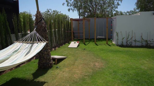 A wide, panning shot of a villa's backyard. The garden features a custom wooden climbing frame with a swing, two cornhole boards for lawn games and a hammock suspended between two palm trees.