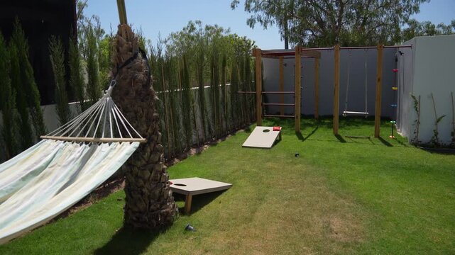  A wide, panning shot of a villa's backyard. The garden features a custom wooden climbing frame with a swing, two cornhole boards for lawn games and a hammock suspended between two palm trees.