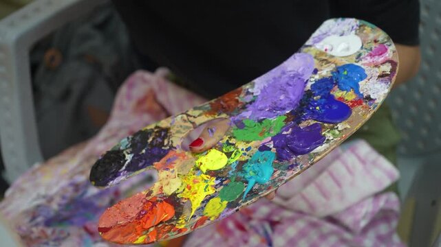 A close-up shot of an artist holding a traditional wooden palette covered in thick, vibrant dabs of colorful paint. The palette shows a messy, authentic mix of textures and hues.
