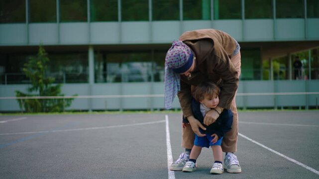 cancer patient zipping toddler jacket outdoors during chemotherapy treatment bending down to care for child showing maternal support parenting responsibility and resilience in daily life