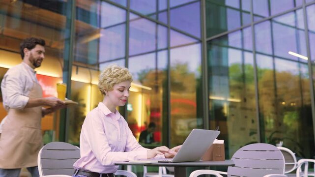 A smiling waiter in an apron serves a fresh salad to a woman sitting at a table with a laptop outside a modern building