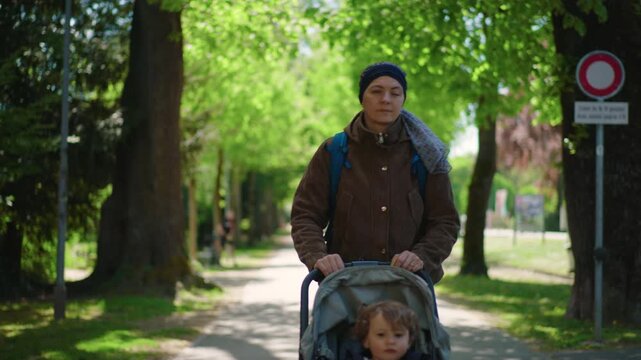 mother with cancer walking forward pushing toddler stroller along tree lined street during chemotherapy treatment showing daily parenting routine resilience and strength while coping with illness