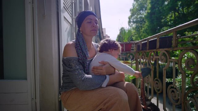 Mother undergoing chemotherapy holding toddler on balcony of residential apartment expressing parental care resilience bonding and quiet urban lifestyle moment