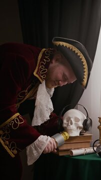 Vertical shot of funny Caucasian man wearing tricorn hat and baroque costume performing comedy song into microphone and playfully handing it to skull model on table