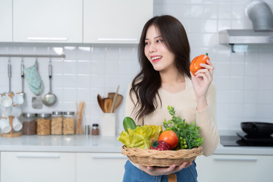 Happy portrait of young asian woman holding a basket of vegetables of standing a cheerful preparing food and enjoy cook cooking with vegetables, while standing on a kitchen Condo life or home