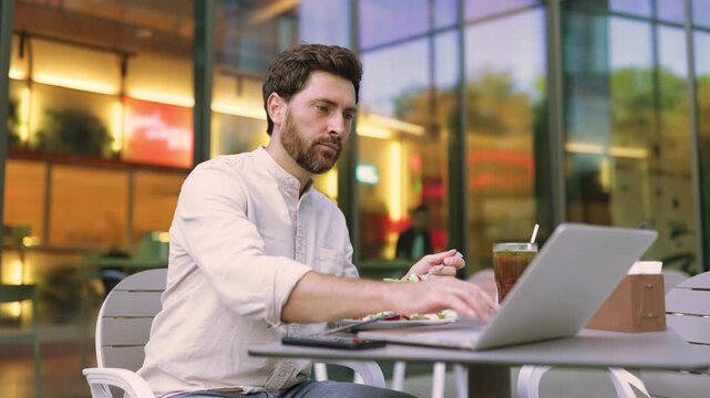 A focused man with a beard works on his laptop while eating a salad at an outdoor cafe with a modern glass building in the background