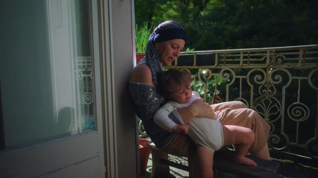 Mother undergoing chemotherapy sitting on balcony holding restless toddler in sunlight expressing caregiving challenge resilience emotional connection and everyday family life during treatment period