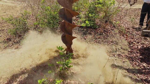 Close-up of a mechanical auger drilling into dry soil on farmland, creating a hole for installing concrete poles and barbed wire fencing, with dust rising in sunlight.