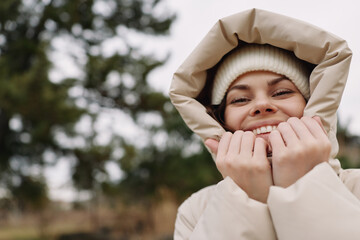 Winter outdoor portrait of a smiling woman in a hooded coat, beanie and warm hands, cozy mood in a chilly natural setting with trees © SHOTPRIME STUDIO