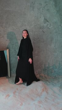 Young beautiful woman in black arabic national dress abaya walking barefoot around chandelier on mirror background