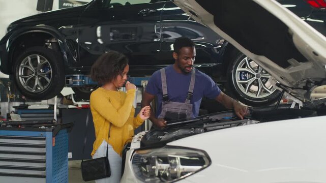 Mechanic explaining a car's breakdown to a female customer, pointing out vehicle parts under the hood to help her understand the problem.