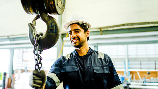 Professional male industrial worker in blue overalls and a white hard hat operates a remote-controlled overhead crane hoist in a factory workshop, looking up at the heavy metal hook and chain.