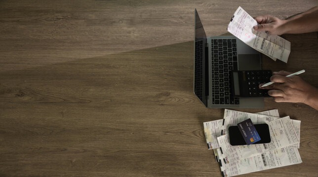 Hands using calculator on a laptop with receipts and credit card on a wooden desk, managing personal expenses and budgeting in a home setting and tax preparation content.