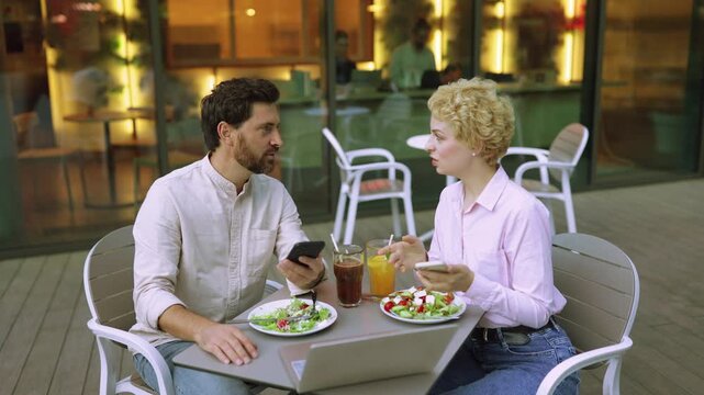 A man and woman are seated at an outdoor cafe table, each looking at their mobile phones while enjoying salads and drinks