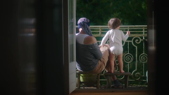 Mother sitting on balcony with toddler standing on small stool then sitting down expressing bonding, caregiving, calm interaction and everyday family life at home