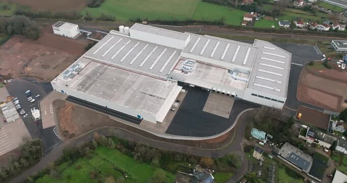 Wide aerial view of a white industrial warehouse facility situated in a green valley with a small village