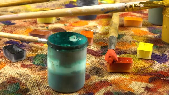 Jars with multi-colored paints, brushes are laid out on a beige burlap cloth with spots of different shapes and colors from gouache and acrylic paints, background, side view