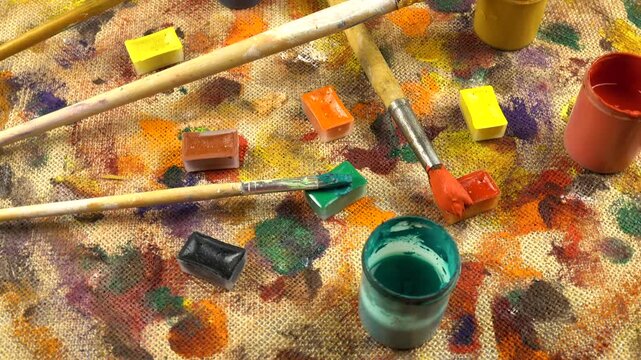 Jars with multi-colored paints, brushes are laid out on a beige burlap cloth with stains of different shapes and colors from gouache and acrylic paints, background, top view