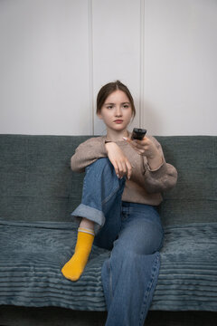 Young girl sitting on a comfortable couch at home, feeling bored and apathetic while navigating channels with a tv remote control, highlighting screen time and leisure indoors. Vertical photo