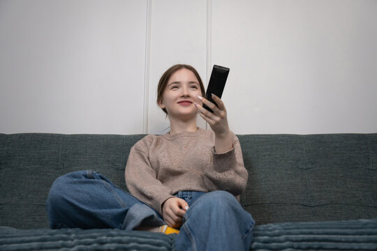 Young girl sitting comfortably on a sofa, holding a remote control and watching television, enjoying leisure time and entertainment in her living room