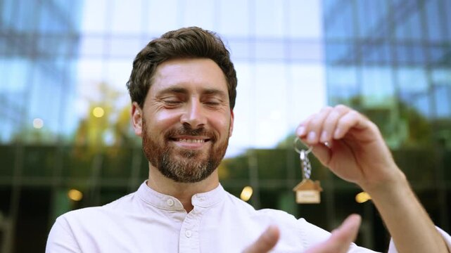 A smiling man proudly holds a set of keys with a small wooden house charm, symbolizing homeownership or a new property