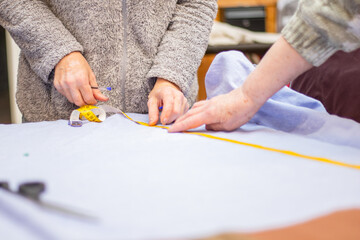 Obraz premium Two hands of individuals measuring fabric with a yellow measuring tape on a light blue textile surface in a sewing cafe environment with sewing tools nearby