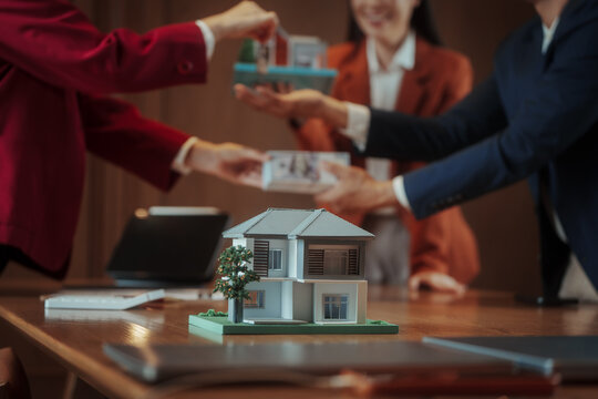 Close-up of hands exchanging keys and money over a table with model house, real estate purchase agreement meeting, agents and buyers in background, warm indoor office.