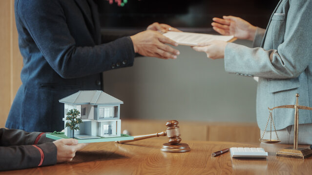 Real estate contract signing with lawyer: hands exchanging documents over desk with model house, gavel and scales of justice, property sale or mortgage closing concept.