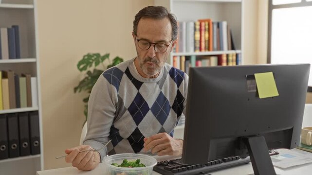Man eating salad at computer in building, fork raised to mouth, hands visible, wearing glasses, desktop monitor, keyboard and mug on desk; focused productivity.