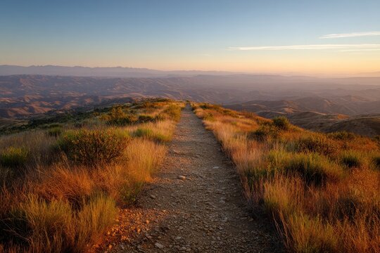 A narrow dirt path leads over a grassy ridge toward a hazy horizon at sunset
