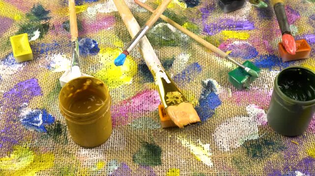 Jars with multi-colored paints, brushes are laid out on a beige burlap cloth with spots of different shapes and colors from gouache and acrylic paints, background, top view