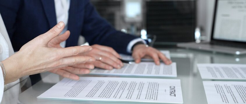 Business people are discussing a contract, reviewing documents and pointing with a pen, sitting at an office table with a laptop, focusing on agreement details and legal paperwork