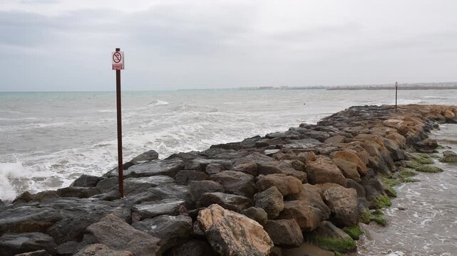 Stormy sea waves hitting rocky breakwater with warning sign