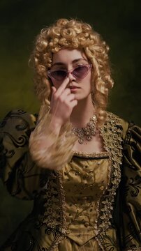 Vertical portrait of confident Caucasian young lady wearing olive embroidered dress, necklace and baroque wig adjusting sunglasses while posing for camera