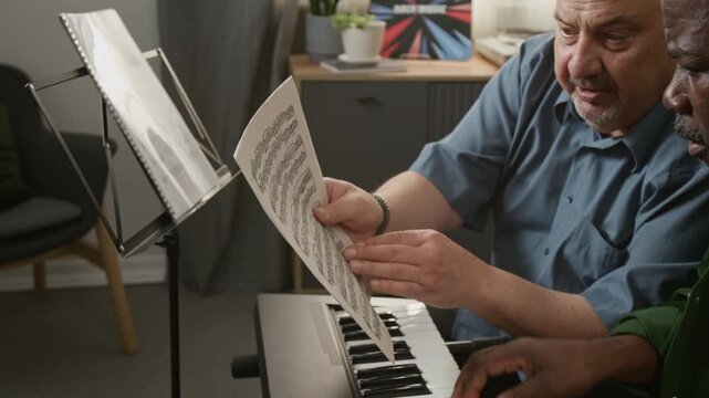 Side view shot of male hands taking note sheet off stand and showing it to senior student learning to play keyboard during music class at home Great for teaching materials or lesson structure examples