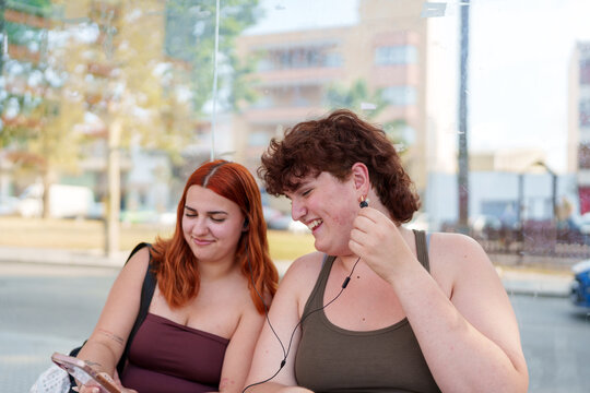 Young women friends sharing wired earbuds and laughing, enjoying music and social media at a bus stop