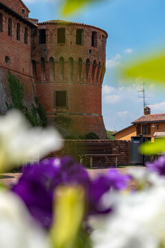 Bologna, Italy - 26 March 2022: View of the imposing red brick walls and rounded tower of Castello di Galeazza, framed by the soft blur of vibrant purple and white flowers in the foreground.