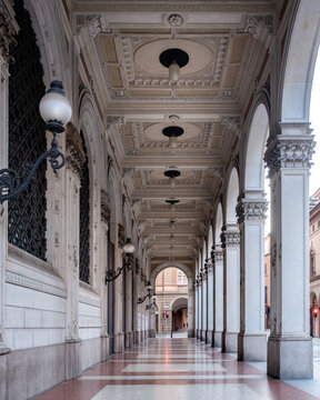 Bologna, Italy - 27 March 2022: View of the porticoes, where the white columns rise to meet the ornately decorated ceiling, casting shadows on the red and white tiled floor.